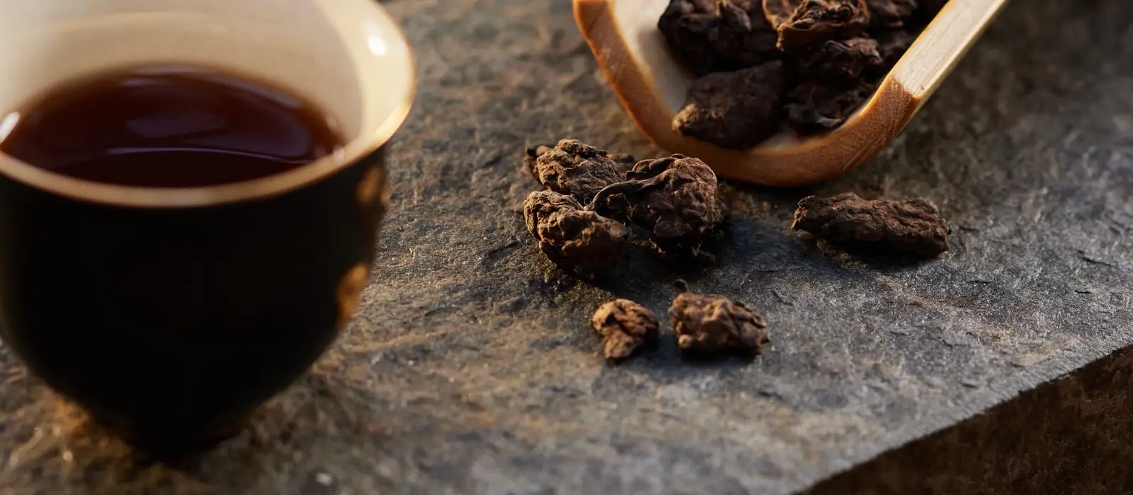 石面茶席上的茶盏与木勺,散落的茶叶在暖光下呈现深浅不一的棕色